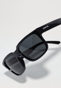 Gafas de sol de plástico negro con lentes de color gris oscuro. Montura de forma cuadrada, textura suave. Nombre de la marca "ARNETTE" impreso en una de las patas.