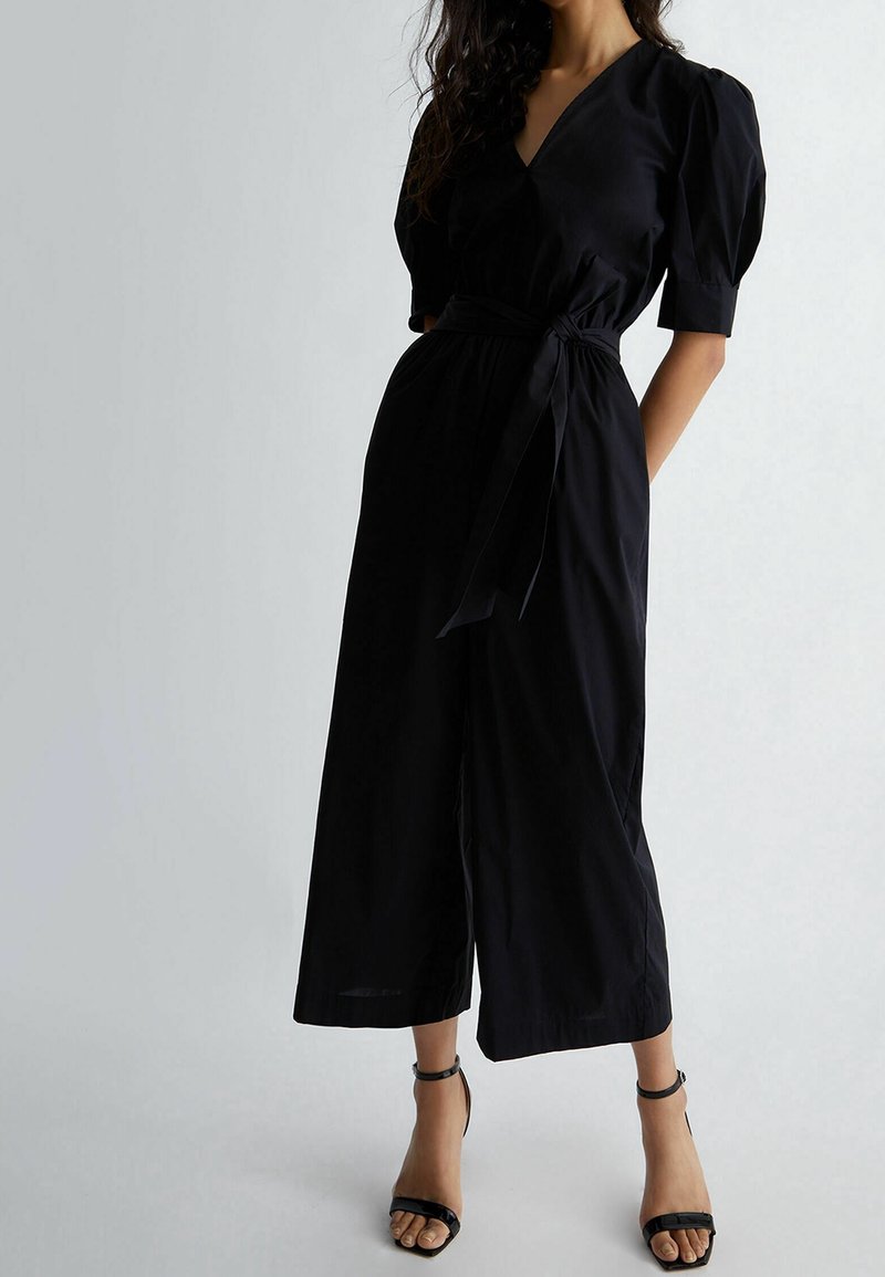 Svart jumpsuit med V-ringning, kort puffärm, midjebälte och vida ben. Bärs med svarta remsandaler med klack. Mjukt tyg.