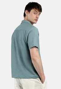Camicia a maniche corte in tessuto testurizzato di un teal sbiadito, con colletto dritto e dettaglio sul giromanica posteriore. Il modello è in piedi con il corpo ruotato e un tatuaggio visibile.