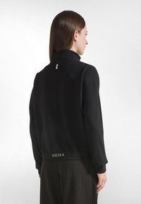 Donna con capelli castani lisci che indossa una giacca nera a maniche lunghe e pantaloni a righe, vista da dietro su uno sfondo bianco.