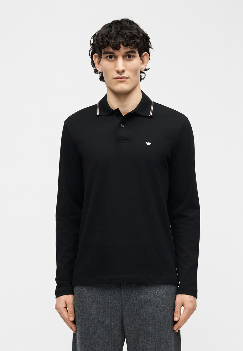 Άνδρας με σκούρα σγουρά μαλλιά, φοράει μαύρο πόλο με μακριά μανίκια και μικρό λευκό λογότυπο στο στήθος και γκρι παντελόνια, στέκεται μπροστά σε απλό φόντο.