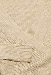 Beige ribbeliger Pullover mit V-Ausschnitt. Das Material zeigt eine weiche Textur mit einem dezenten Glanz. Betonte Kanten und Falten sind sichtbar.