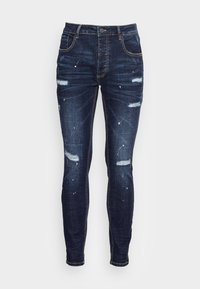 Vaqueros skinny de denim azul oscuro con detalles desgastados, que presentan desgarros, salpicaduras de pintura y costuras en contraste. Diseño estándar de cintura.