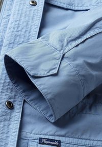 Manica di una camicia a maniche lunghe azzurro chiaro con polsino visibile, bottone a pressione argento e etichetta "Façonnable" nella parte inferiore della camicia.