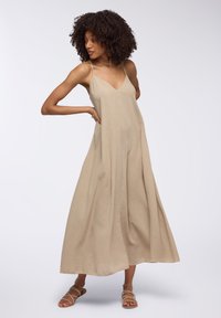 Abito maxi beige senza maniche con scollo a V, silhouette fluida e tessuto texturizzato. Abbinato a sandali con cinturini.