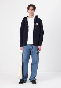 Veste à capuche zippée navy avec cordons blancs sur un t-shirt blanc à col rond, associée à un jean cargo bleu clair et des chaussures noires, portant un sac fourre-tout beige.