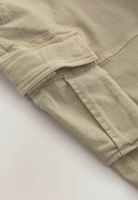 Gros plan d'une manche en tissu beige avec des coutures détaillées et une sangle en tissu fixée par un rabat avec des coutures visibles.