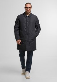 Zwarte geïsoleerde jas met een knopensluiting, een gespreid kraag, zijzakken en ribgebreide manchetten; gedragen over donkere broeken en sneakers.