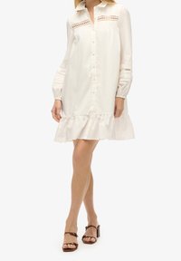 Vestito in cotone bianco con bottoni, caratterizzato da un orlo arricciato, maniche lunghe con dettagli in pizzo e colletto classico. Texture liscia, vestibilità comoda.