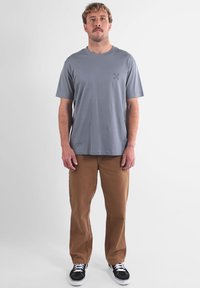 T-shirt en coton gris à manches courtes et col rond, associé à un pantalon marron et des baskets noires. Design minimaliste avec un logo à l'avant.