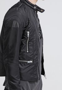 Svart bomberjacka med en figursydd design, som har kviltade axeldetaljer, flera silverfärgade dragkedjor och en slät, glänsande tygtextur.