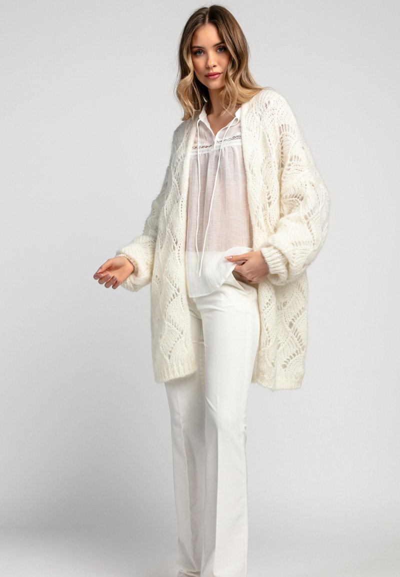 Biała dziergana kardigan z koronkowym wzorem, w oversize'owym kroju, z długimi rękawami, noszony na białej bluzce i zestawiony z białymi spodniami.