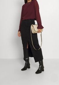 Schwarzer Jeansrock mit einem Schlitz vorne, kombiniert mit einer burgunderfarbenen, strukturierten Bluse. Accessoires sind eine hellbeige Umhängetasche und schwarze Stiefel mit Absatz.