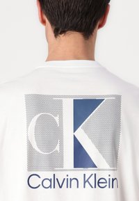 T-shirt blanc à manches courtes avec un grand graphique au dos présentant un "CK" bleu marine et "Calvin Klein" en bleu sous le design.