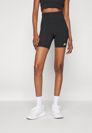 Woman wearing black high-waist athletic shorts with Adidas logo, white crew socks, and white athletic shoes, standing against a plain background.