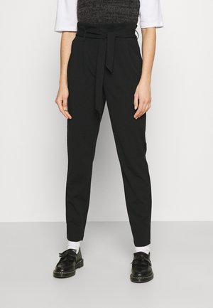 Black high-waisted tapered pants with a tied belt worn with white socks and black tassel loafers on a person standing against a white wall.