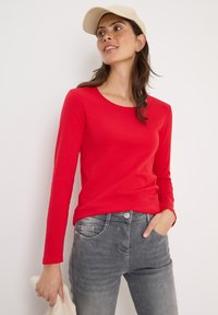 Röd långärmad tröja med rund halsringning i kombination med grå slitna jeans. Personen håller ett ljusfärgat föremål i handen.