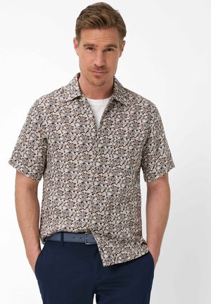 Mand iført en beige og sort kortærmet blomstret skjorte over en hvid undertrøje, stoppet ned i mørkeblå bukser med et matchende bælte.