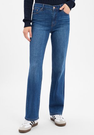 Person trägt gerade geschnittene blaue Jeans, weiße Sneakers mit schwarzen Streifen und einen dunklen Langarmpullover, steht vor einem einfarbigen Hintergrund.