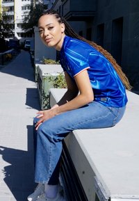 Blå sporttröja med grafisk design, vita accenter och korta ärmar. Kombinerad med ljusblå denimjeans och vita sneakers. Utomhusmiljö.