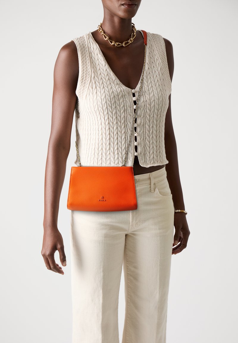 Borsa a tracolla arancione in pelle con texture liscia e tracolla in argento, indossata sopra un top a maglia crema e pantaloni bianchi.