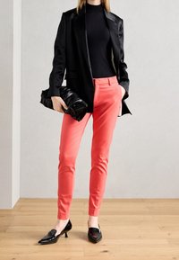 Zwarte blazer over een zwarte top, gecombineerd met fel koraalkleurige broek. Accessoires zijn onder andere een gestructureerde zwarte clutch en zwart spitse schoenen.