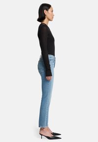 Schwarzes Langarmoberteil, hellblaue High-Waist-Jeans mit ausgefransten Säumen und schwarze spitze Absätze; Modell im Profil dargestellt.