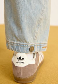 Rudos zomšo Adidas sportbačiai su kreminiu kulno užsegimu ir juodu logotipu, iš dalies uždengti šviesiai mėlynomis džinsinėmis kelnėmis su metaliniu mygtuko detalumu.