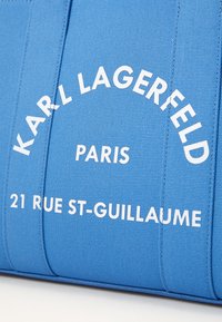 Niebieska torba z płótna z białym napisem "KARL LAGERFELD PARIS 21 RUE ST-GUILLAUME." Napis jest pogrubiony i wyśrodkowany.