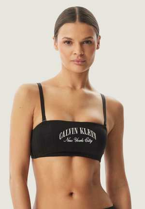 Femme aux cheveux bruns et raides porte un haut bandeau noir Calvin Klein New York City avec fines bretelles sur un fond clair uni.