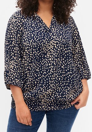 Vrouw die een marineblauwe blouse draagt met een beige onregelmatig stippenpatroon en driekwartmouwen, gecombineerd met een blauwe spijkerbroek.