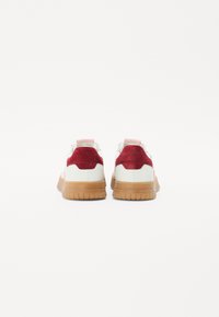 Tenisice z belim usnjem, rdečim semiš petam in poudarki, kontrastnim gumijastim podplatom, zaobljeno obliko in zanko na hrbtni strani.
