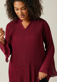Haut plissé bordeaux avec un col en V, des manches cloche longues et une finition texturée. Le tissu présente un léger relief et une silhouette fluide.