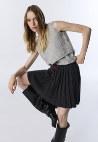 Jeune femme portant un haut en tricot sans manches gris, une jupe plissée sombre avec un cordon rouge, et des bottes noires jusqu'aux genoux dans une pose dynamique.