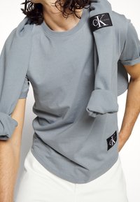 Persona vistiendo una camiseta de Calvin Klein color azul claro y un suéter a juego colocado sobre los hombros, con pantalones blancos, en una pose en medio de una acción.