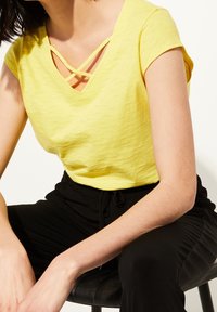Žlutý krátký rukáv V-čelní top s křížovým.Detailing straps na výstřihu, vyrobený z texturované látky. Nosit s černými kalhotami na elastický pas.