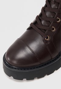 Bottine en cuir marron avec un bout rond, design à lacets, œillets en doré et semelle plateforme noire épaisse. Fini texturé avec des détails de couture.