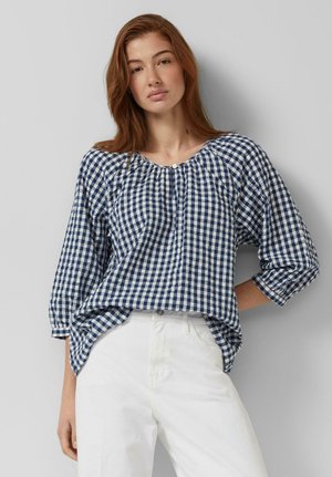 Jonge vrouw met lang rood haar, gekleed in een blauw-wit geruite blouse en witte broek, staand tegen een effen grijze achtergrond.