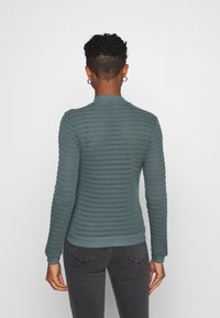 Gebreide trui in petrolblauw met een gestructureerd patroon, voorzien van een ronde halslijn en geribde manchetten. Achteraanzicht toont een aansluitend ontwerp.