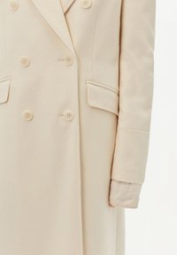 Doppelreihiger cremafarbener Mantel mit weicher Textur, großen Knöpfen, zwei vorderen Taschen und taillierter Silhouette.