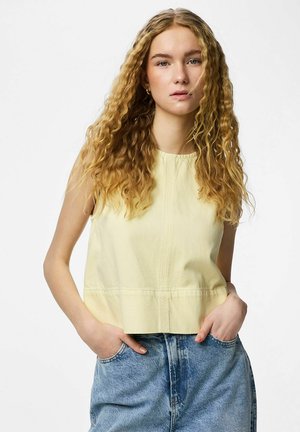 Jeune femme aux longs cheveux blonds bouclés, portant un haut sans manches jaune pâle et un jean en denim bleu, les mains dans les poches, expression neutre.