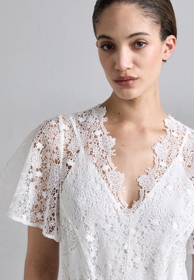 Witte jurk met ingewikkelde kantdetails, een V-halslijn en korte, fladderende mouwen met scalloped randen, wat een delicate textuur geeft.