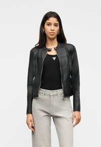 Νεαρή γυναίκα που φοράει μαύρο τζάκετ με φερμουάρ, μαύρο τοπ και ανοιχτό γκρι τζιν, στέκεται μπροστά σε απλό ανοιχτό φόντο.