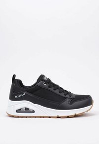 Zapatillas deportivas negras con textura de malla y lisa, suela blanca, detalle de amortiguación de aire y diseño con cordones. Presenta un logo en el lateral.