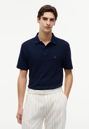 Jeune homme portant un polo bleu marine texturé rentré dans un pantalon blanc à fines rayures avec une ceinture blanche, debout avec une main dans la poche.