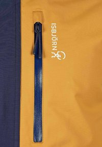 Blå lynlås på sennepsgul stof ved siden af et marineblåt stofpanel, med hvid tekst "ISBJÖRN" og et lille logo printet ved siden af lynlåsen.