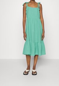 Une robe mi-longue vert menthe avec un décolleté carré, des bretelles nouées aux épaules et une jupe à volants froncés. Portée avec des sandales blanches et noires.