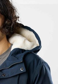 Person with curly hair wearing a navy blue jacket with a white fleece-lined hood and a gray shirt underneath.