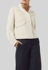 Kremowa kurtka z wysokim kołnierzem, dwoma kieszeniami na piersi i widocznymi przeszyciami. Połączona z ciemnymi dżinsowymi spodniami ozdobionymi złotymi guzikami.