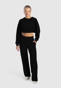 Schwarzes, verkürztes Sweatshirt und locker sitzende schwarze Hose, beide aus weichem Stoff. Das Sweatshirt hat einen runden Ausschnitt; die Hose hat einen elastischen Bund.